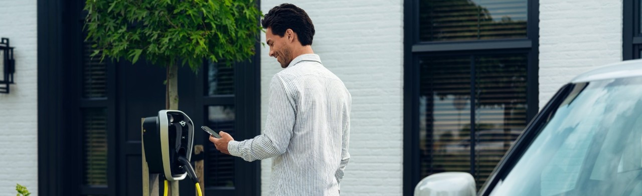 Man looks at his phone while standing in front of a home charging station with a blue MINI behind him.