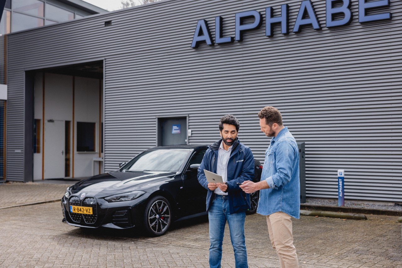 Two people standing next to a car, discussing information displayed on a tablet in an outdoor setting.
