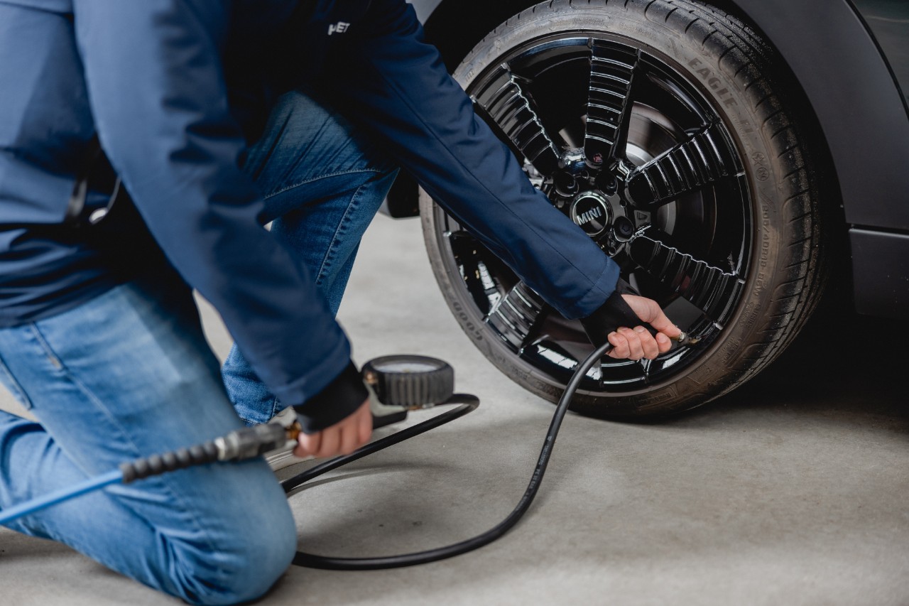 A person wearing an Alphabet jacket checks and inflates the tyre of a car in a well-lit underground car park.