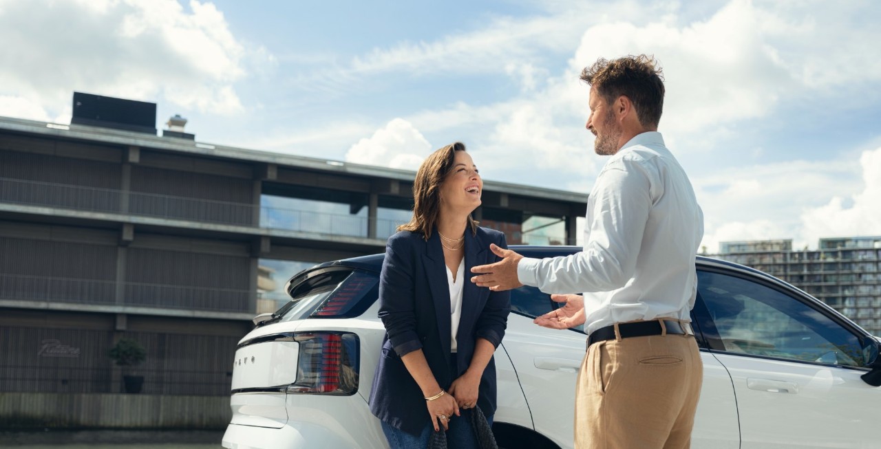 A woman leans against a white car and has a conversation with a man.