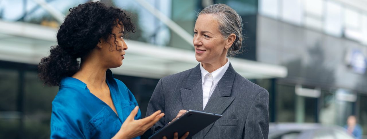 Two women have a conversation in front of a building with large glass windows.