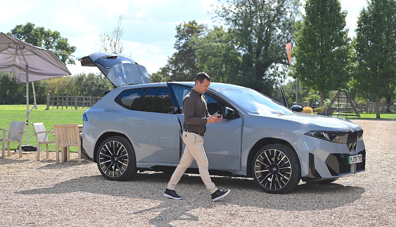 Person walking past a parked grey BMW SUV with the driver door and rear hatch open on a sunny outdoor event site, with chairs and trees in the background.