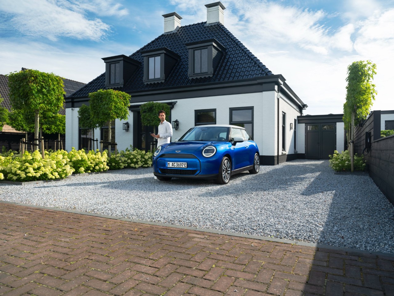 Man stands next to blue mini parked on a gravel drive in front of a modern house.