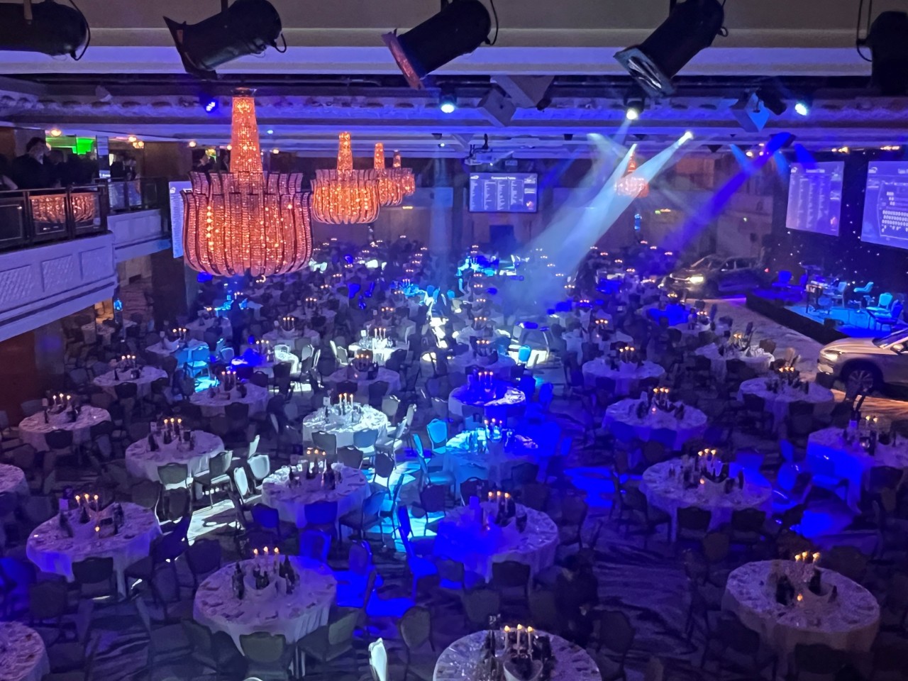 "Large banquet hall set up for an awards event, with round tables covered in white tablecloths and candles, crystal chandeliers hanging from the ceiling, stage with spotlights and large screens, and cars displayed near the stage. The room is illuminated with blue and purple lighting, and people are visible on the balcony.