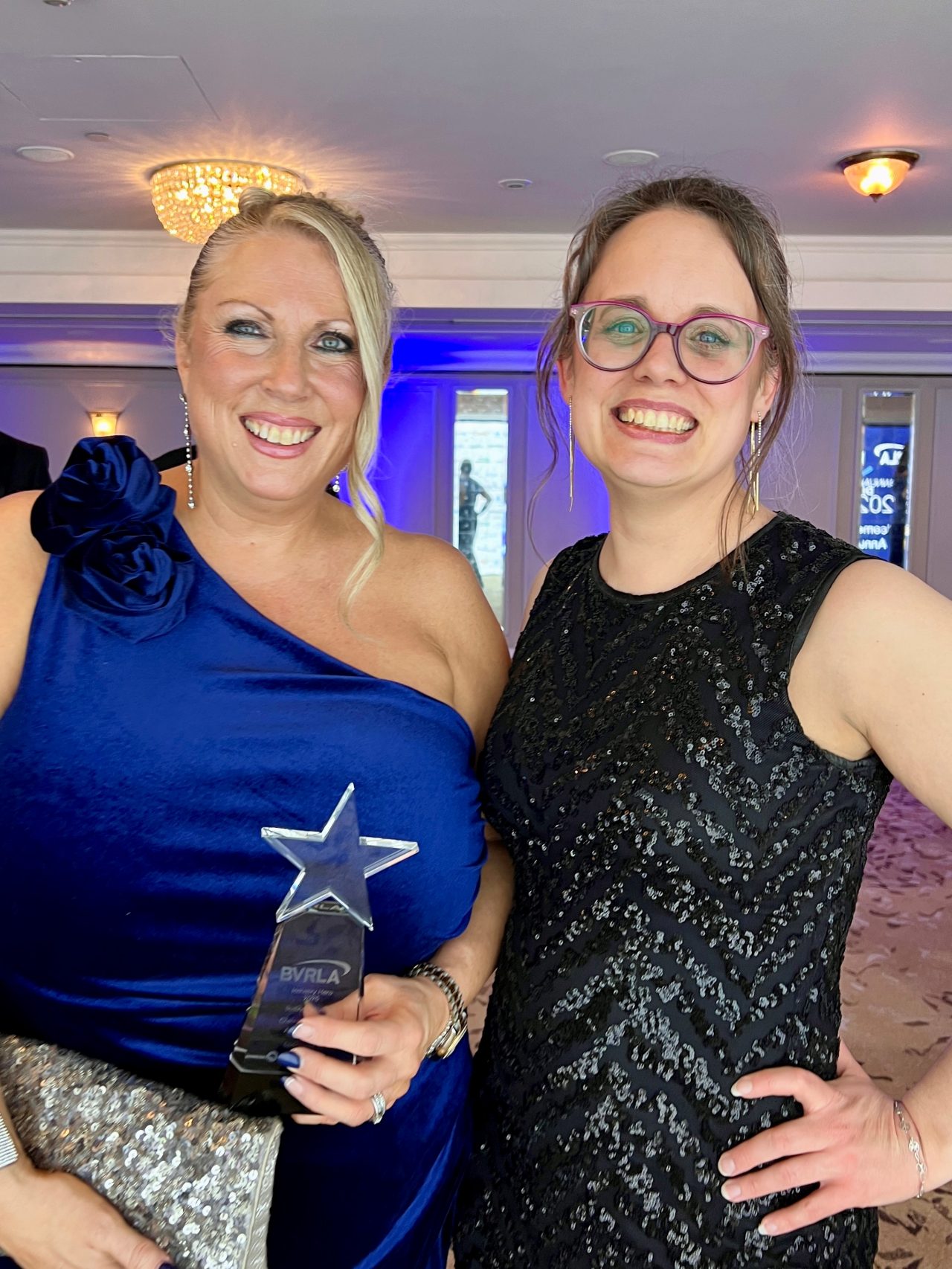Two smiling women posing indoors at an event. The woman on the left wears a dark blue one-shoulder dress with a floral detail, holding a star-shaped award and a sparkly silver clutch. The woman on the right wears a sleeveless black sequined dress and glasses. Chandeliers and purple lighting are visible in the background.