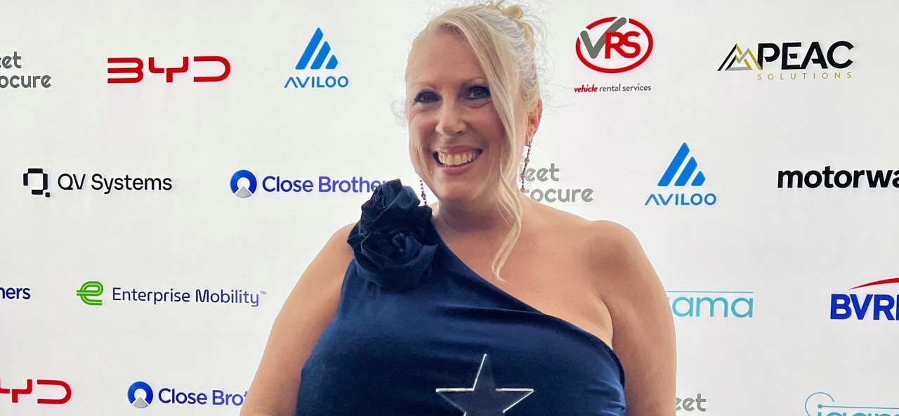 Claire Coomber in a dark blue one-shoulder evening gown holding a star-shaped award and a sparkly silver clutch, standing in front of a backdrop with multiple company logos.