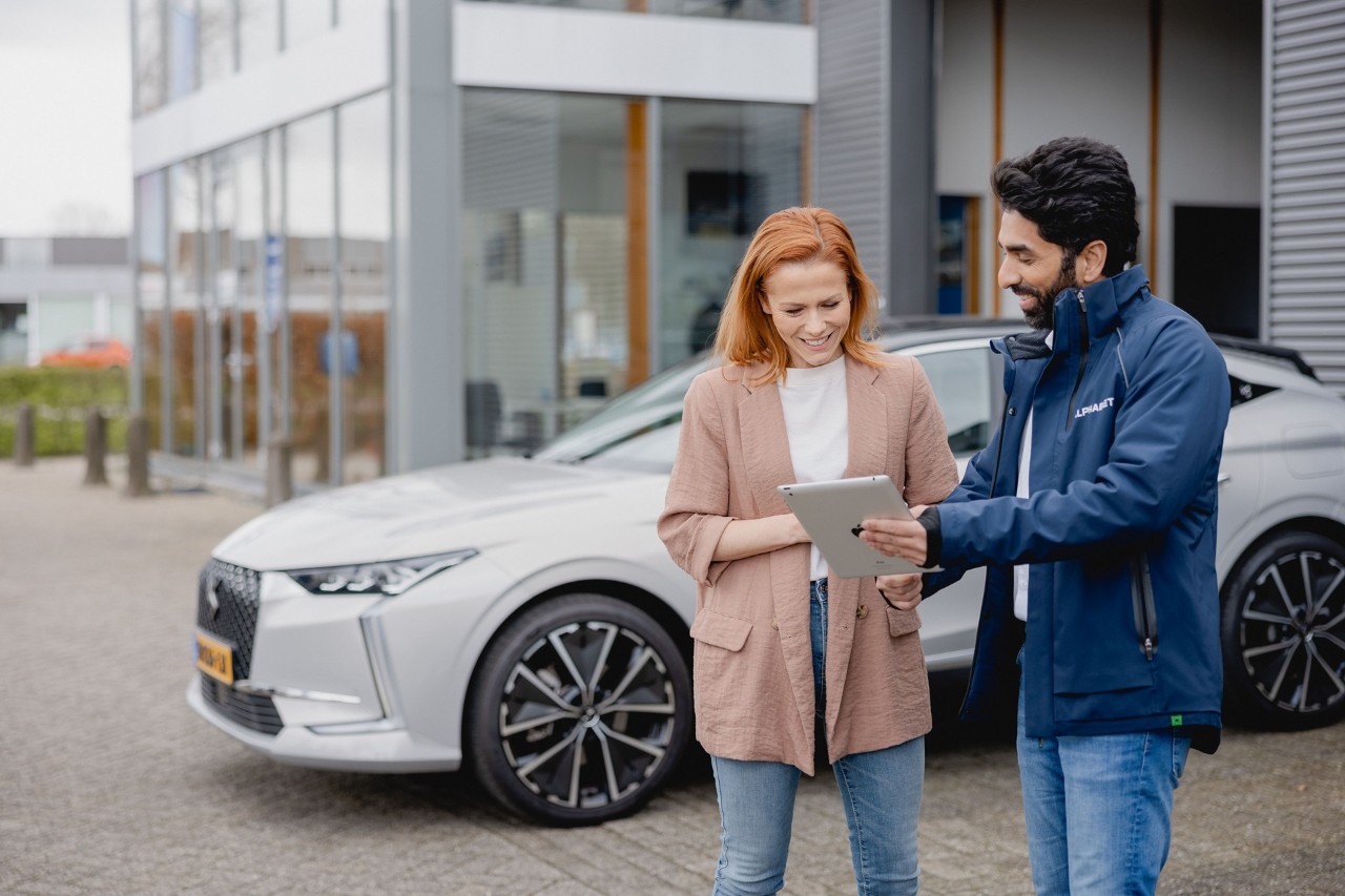 Man and woman in a car dealership, smiling and looking at a tablet outside with a modern car in the background.