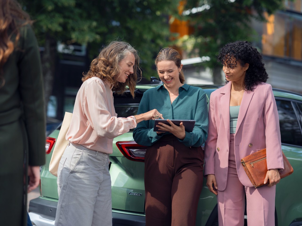 Three women discuss in front of a green car, holding a tablet.