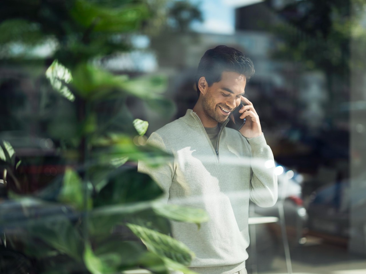 Man smiling on the phone looking down. A plant is next to him in the foreground.