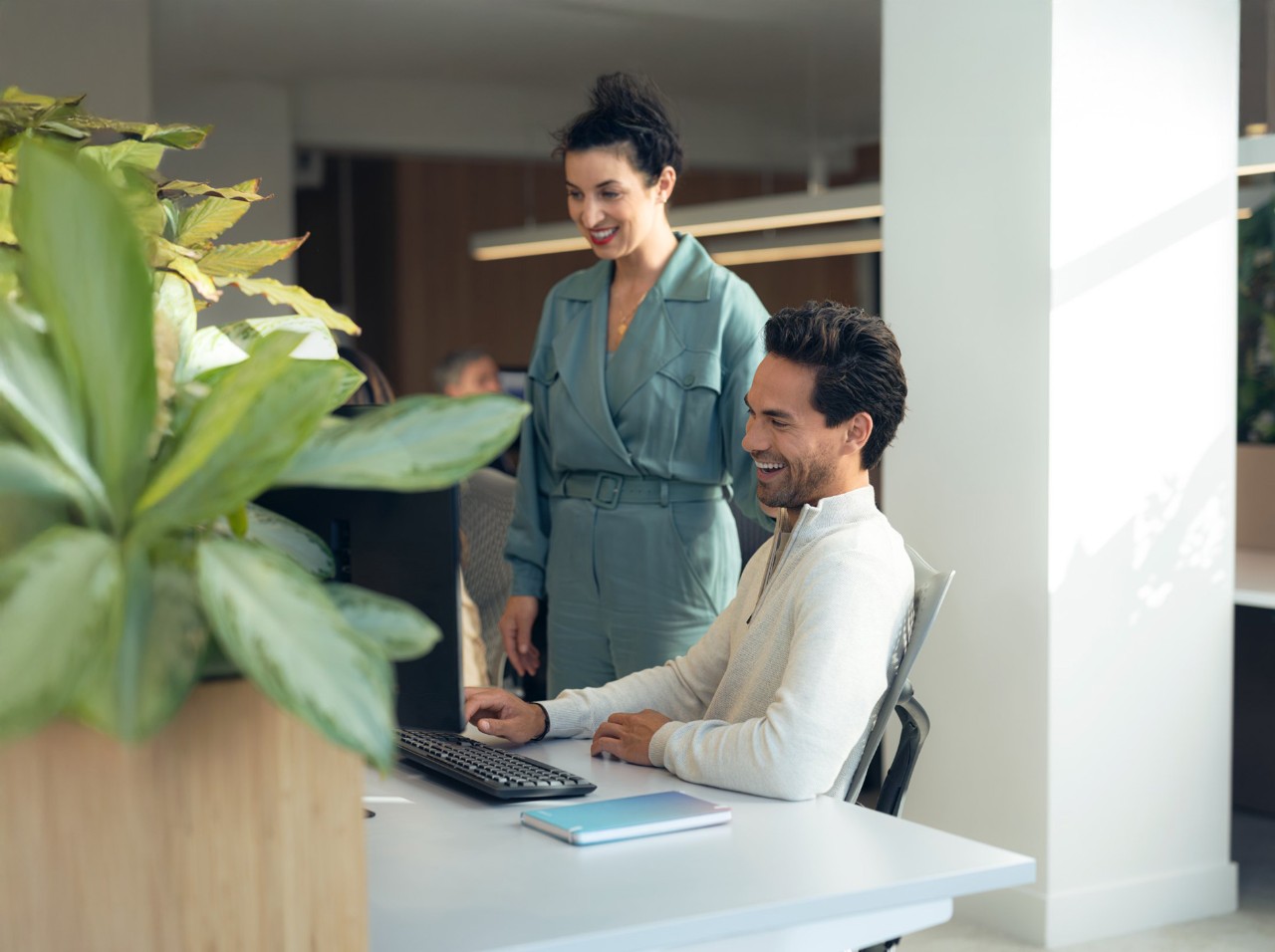 man sitting at a desk, smiling and working on his computer. A woman stands behind him, looking at the screen with a friendly expression. The office has a modern, light-filled atmosphere with plants and colleagues in the background.
