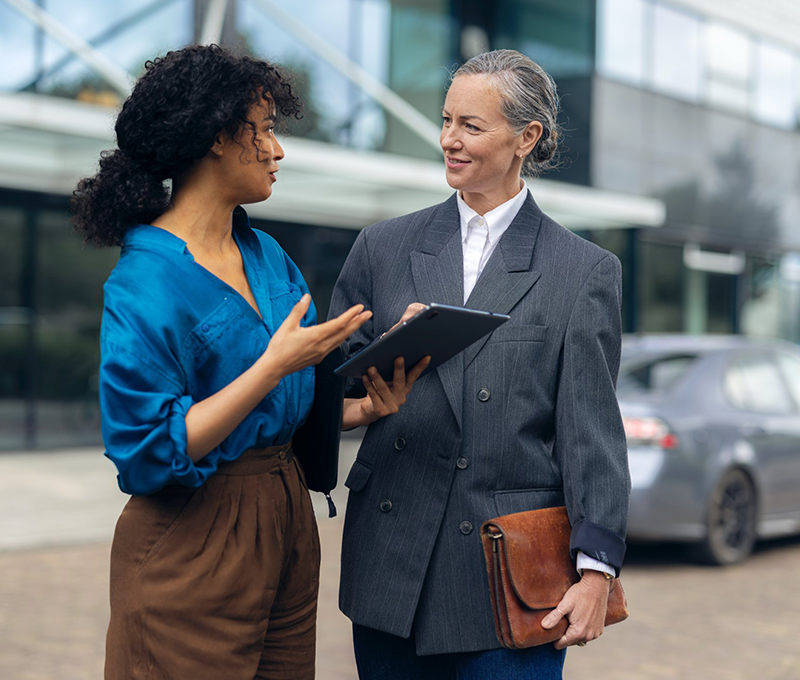 Two women have a conversation in front of a building with large glass windows.