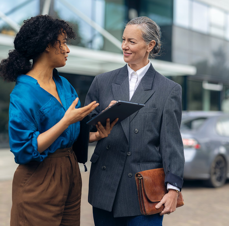 Two women have a conversation in front of a building with large glass windows.