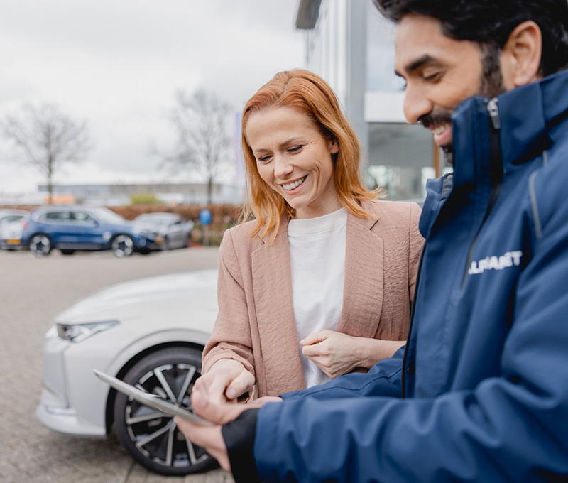 Two people standing next to a white car, discussing information displayed on a tablet in an outdoor setting.