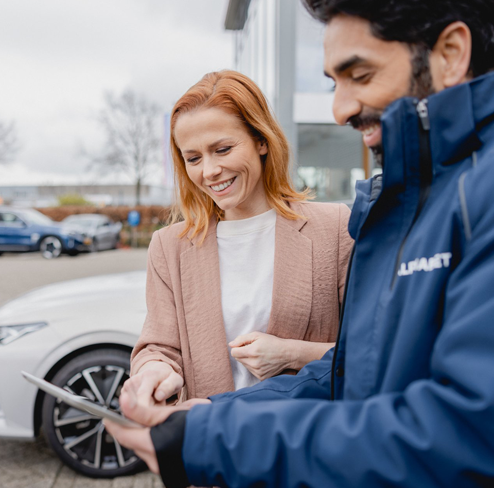Two people standing next to a white car, discussing information displayed on a tablet in an outdoor setting.