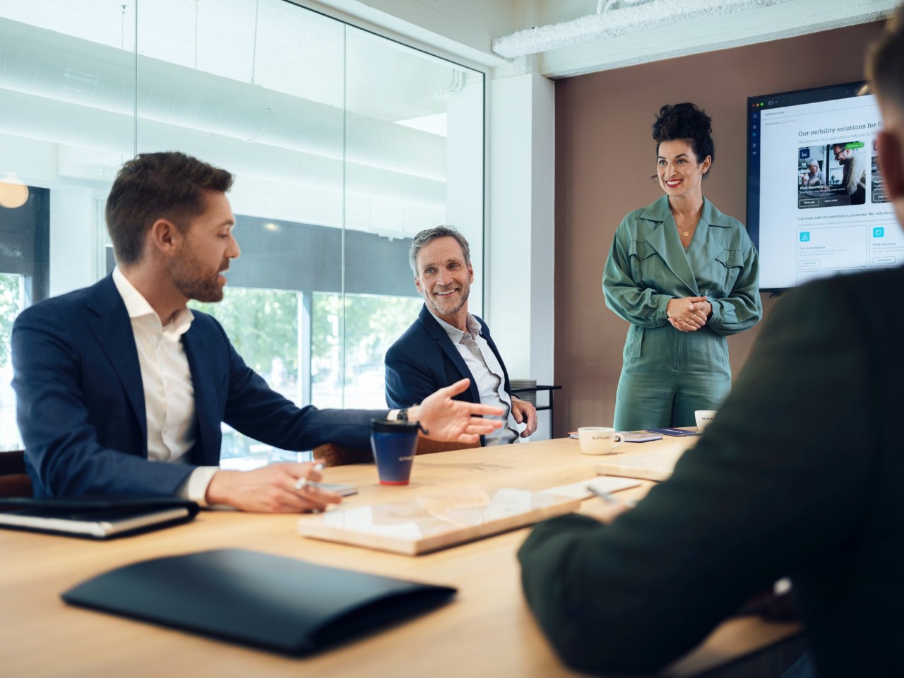 Three people are having a discussion at a wooden table in a modern conference room with a large screen.