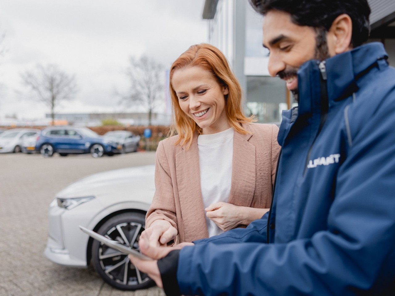 Two people standing next to a white car, discussing information displayed on a tablet in an outdoor setting.