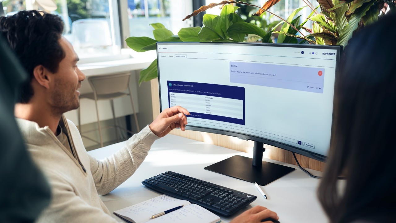 Person sitting at a desk in a modern office, interacting with data on a widescreen monitor, with a notebook and keyboard on the table and green plants in the background.