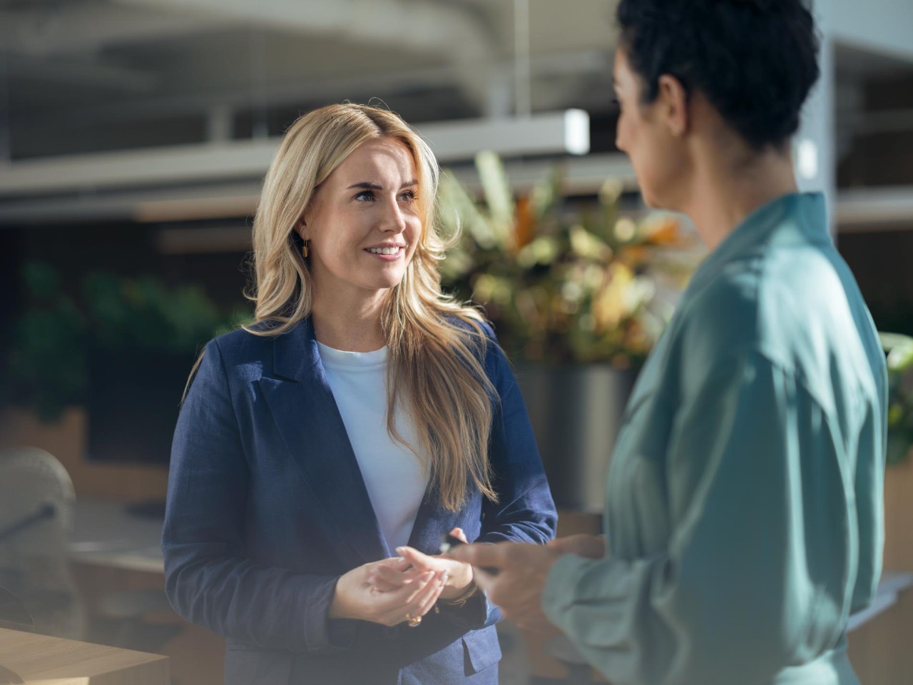 Two women stand facing each other and talking in a modern indoor space with plants; one, wearing a blue blazer and white shirt, smiles while listening as the other gestures during the conversation.