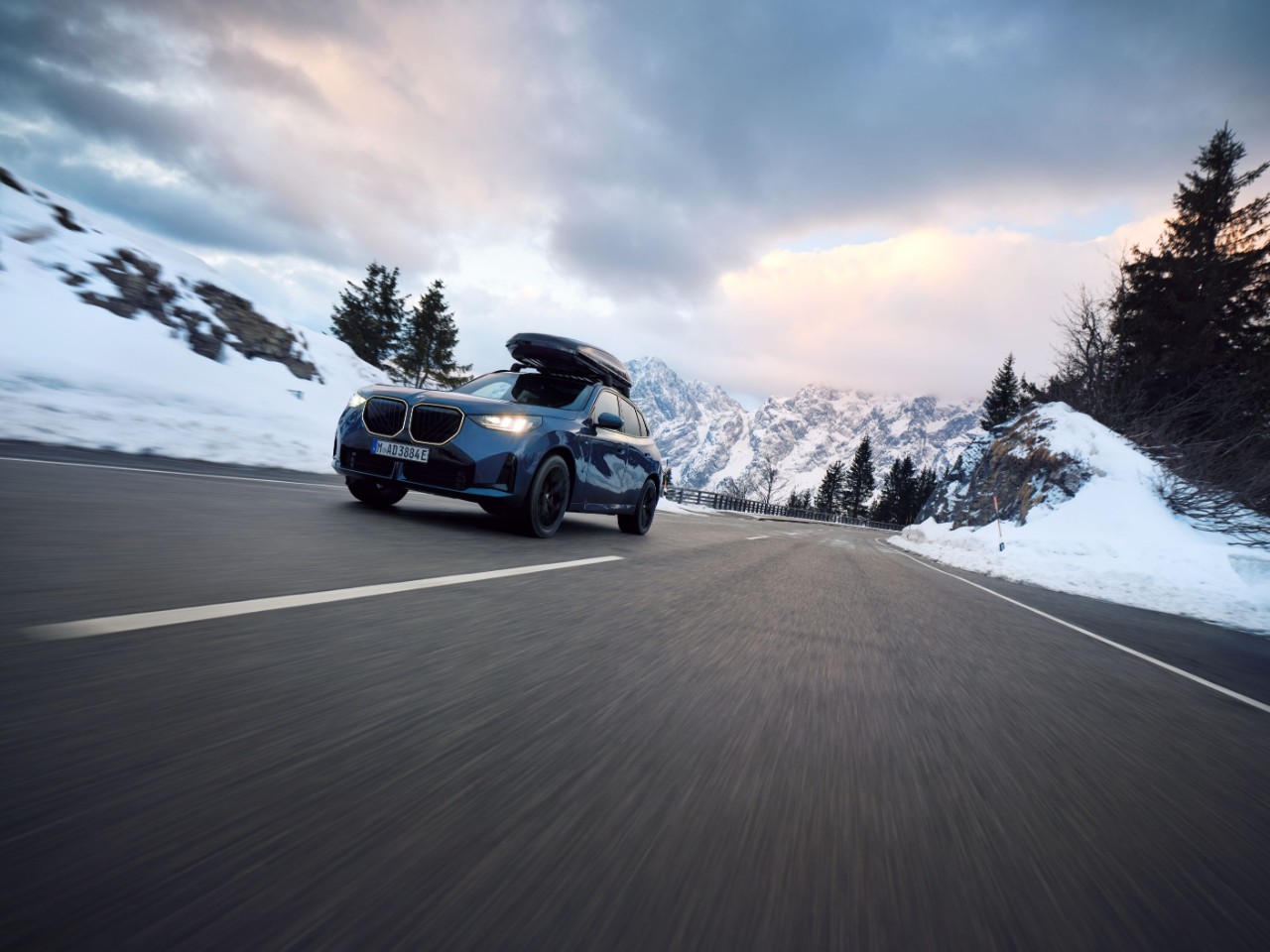 A blue car with a roof cargo box drives on a snowy mountain road at sunset. Snow-covered peaks and evergreen trees line the road, creating a serene winter scene.