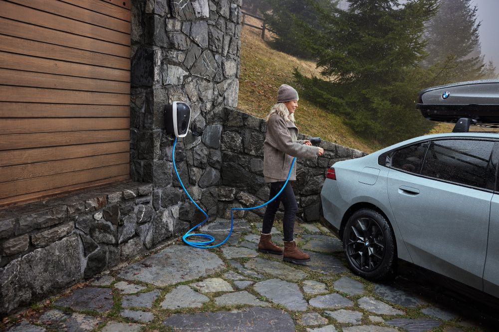 A woman charges an electric car in a rustic, stone-paved driveway surrounded by trees. The setting is calm and eco-friendly.