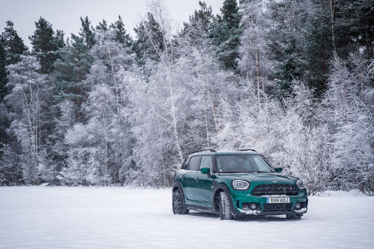 A green car on a snow-covered landscape with frosted trees and a dense forest backdrop. The scene feels serene and chilly, capturing winter's essence.
