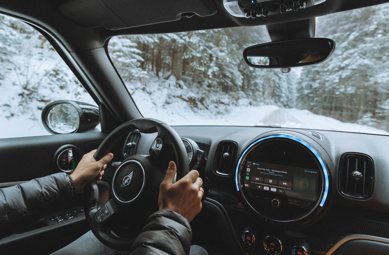 Interior view of a car being driven through a snowy forest. The dashboard displays navigation, and the driver grips the wheel, conveying focus.
