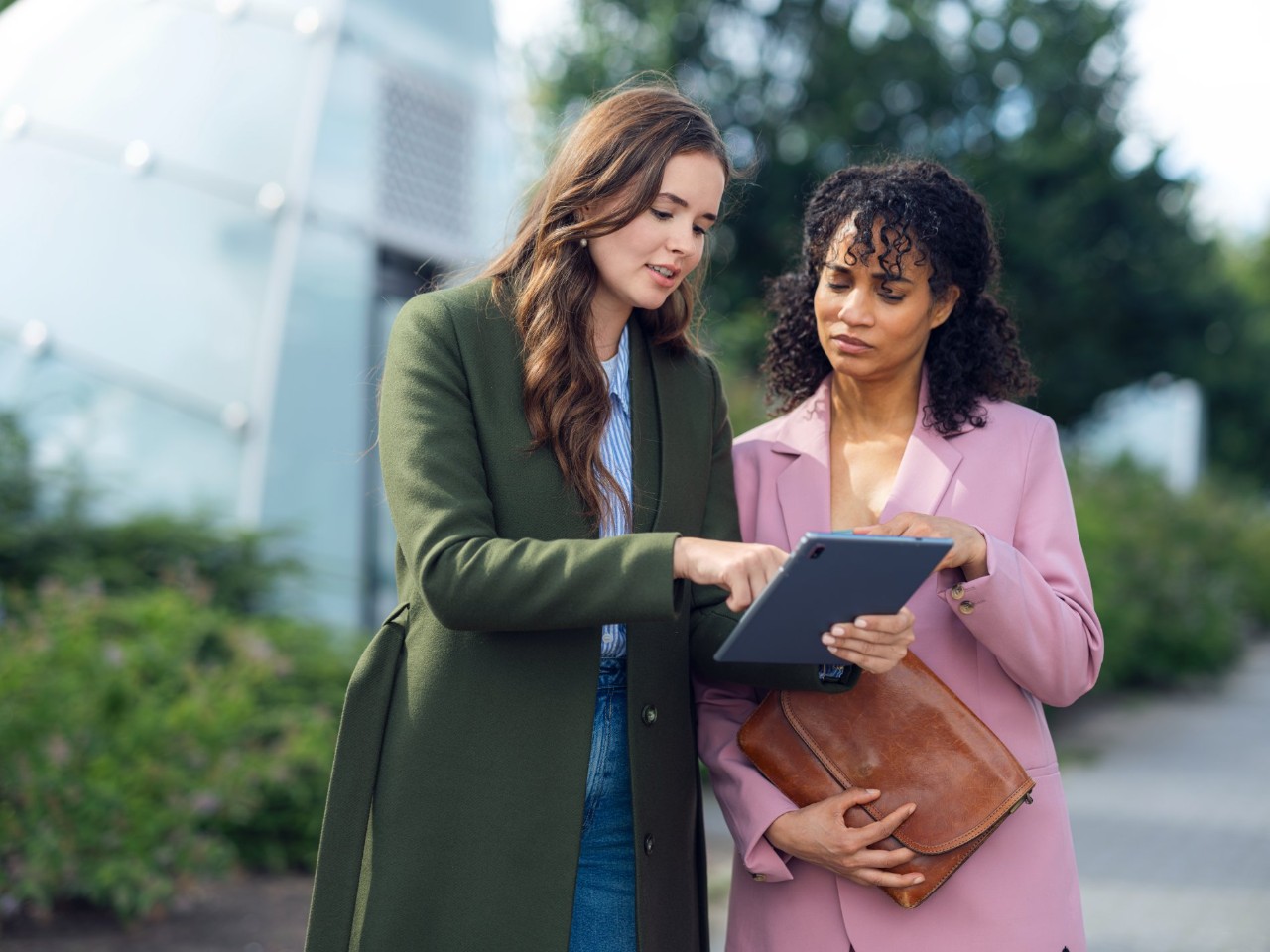 Two women stand on the sidewalk and look intently at a tablet.