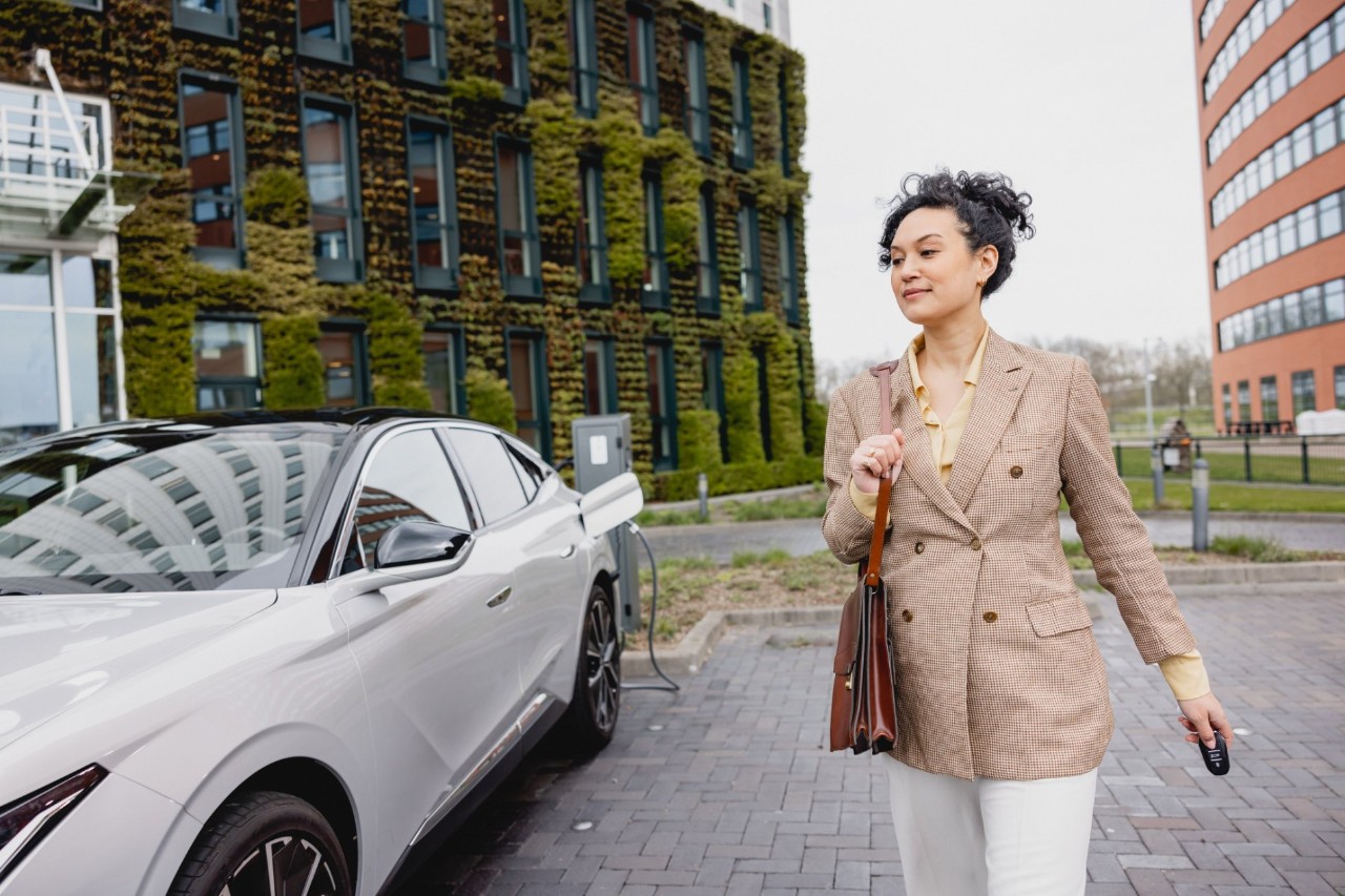 Woman in blaze walks away from electric car in charging port she holds her car keys and a breifcase there is a office building in the background.