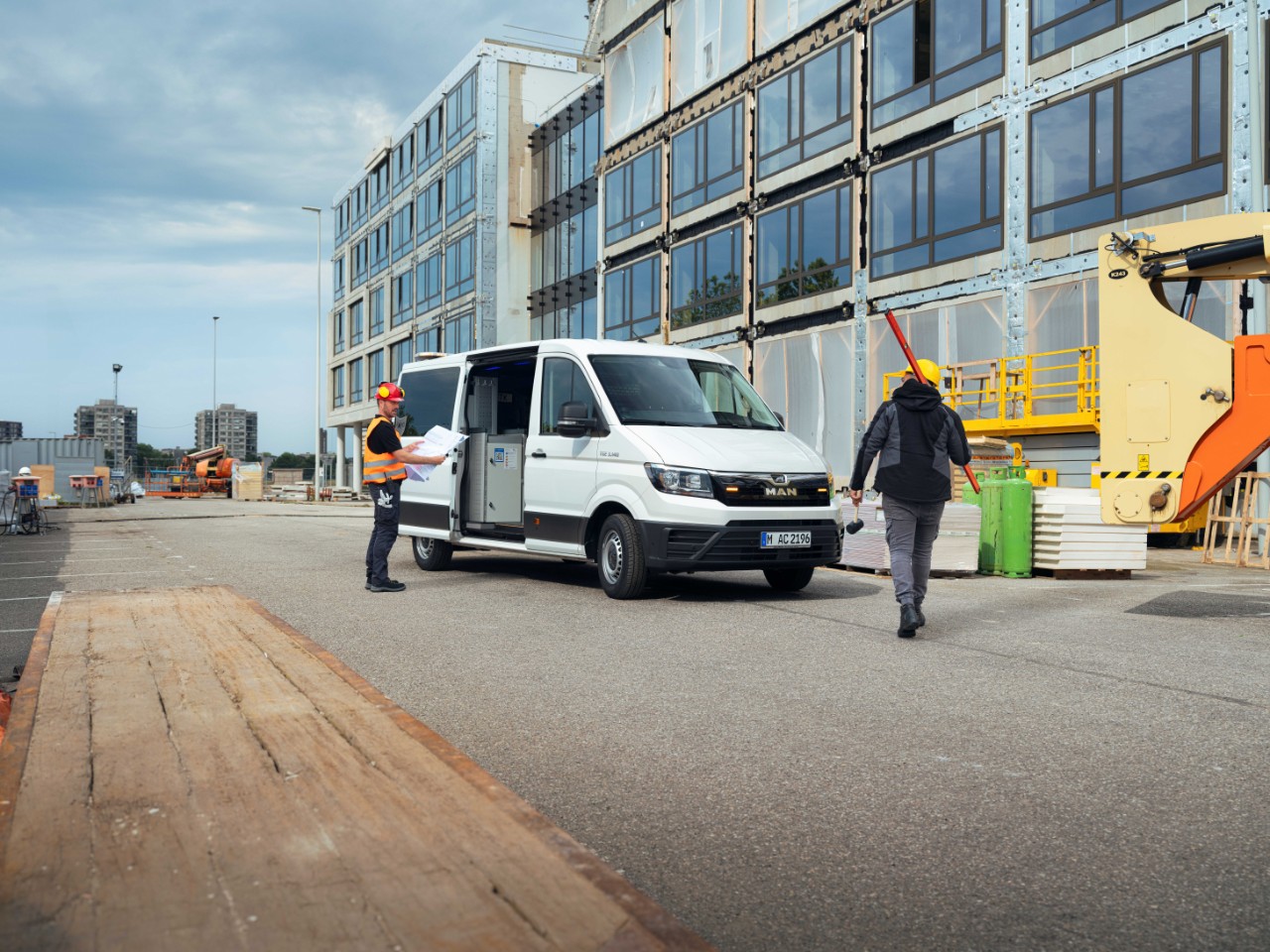 Two construction workers on a building site. One is holding blueprints and standing near a white delivery van, while the other walks away, carrying a tool. The construction site, with a large building under development and machinery in the background, is visible.
