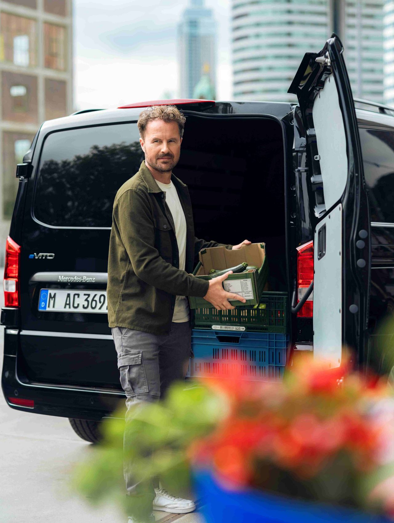 A man unloading a box of produce from the back of a black van. He is standing outdoors with modern city buildings in the background. The van's rear doors are open, and crates of vegetables are visible inside. Colourful flowers are blurred in the foreground.