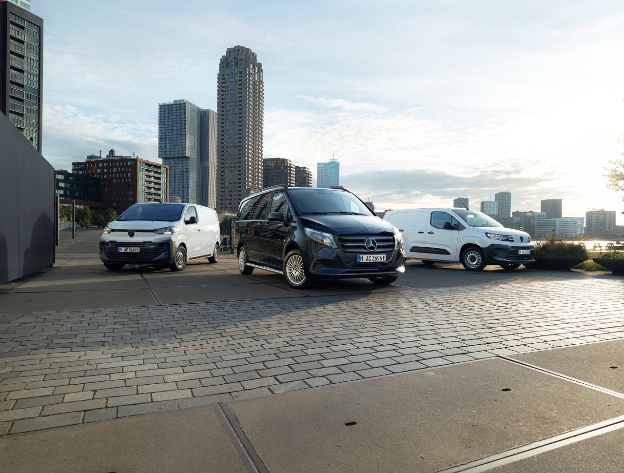 Three vans parked in a city setting with tall buildings in the background. The vans, one black and two white, are arranged in the foreground on a paved surface, with modern urban architecture and a skyline visible in the distance.