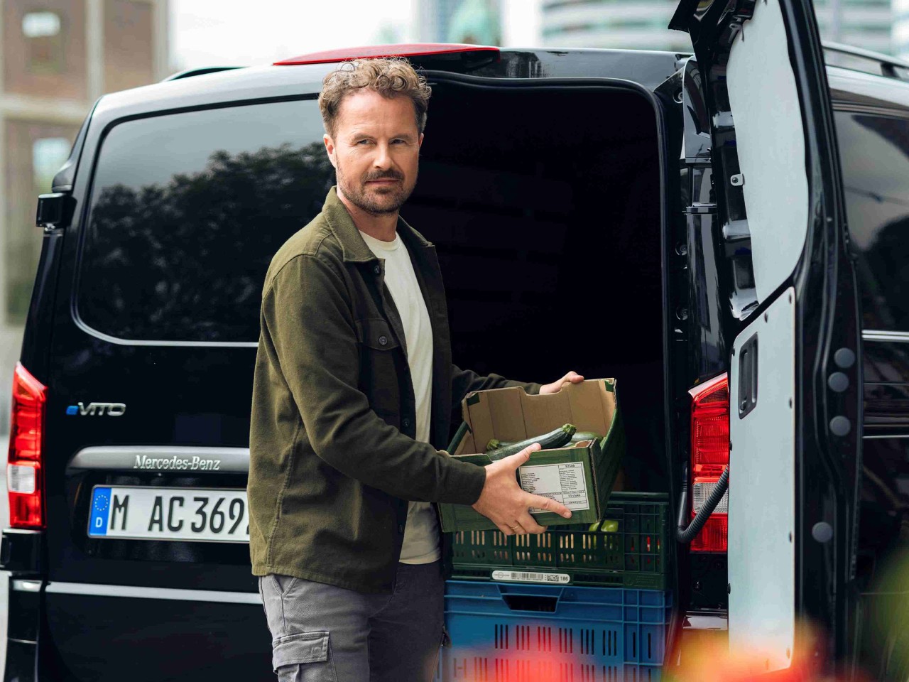 A man unloading a box of produce from the back of a black van. He is standing outdoors with modern city buildings in the background. The van's rear doors are open, and crates of vegetables are visible inside. Colourful flowers are blurred in the foreground.