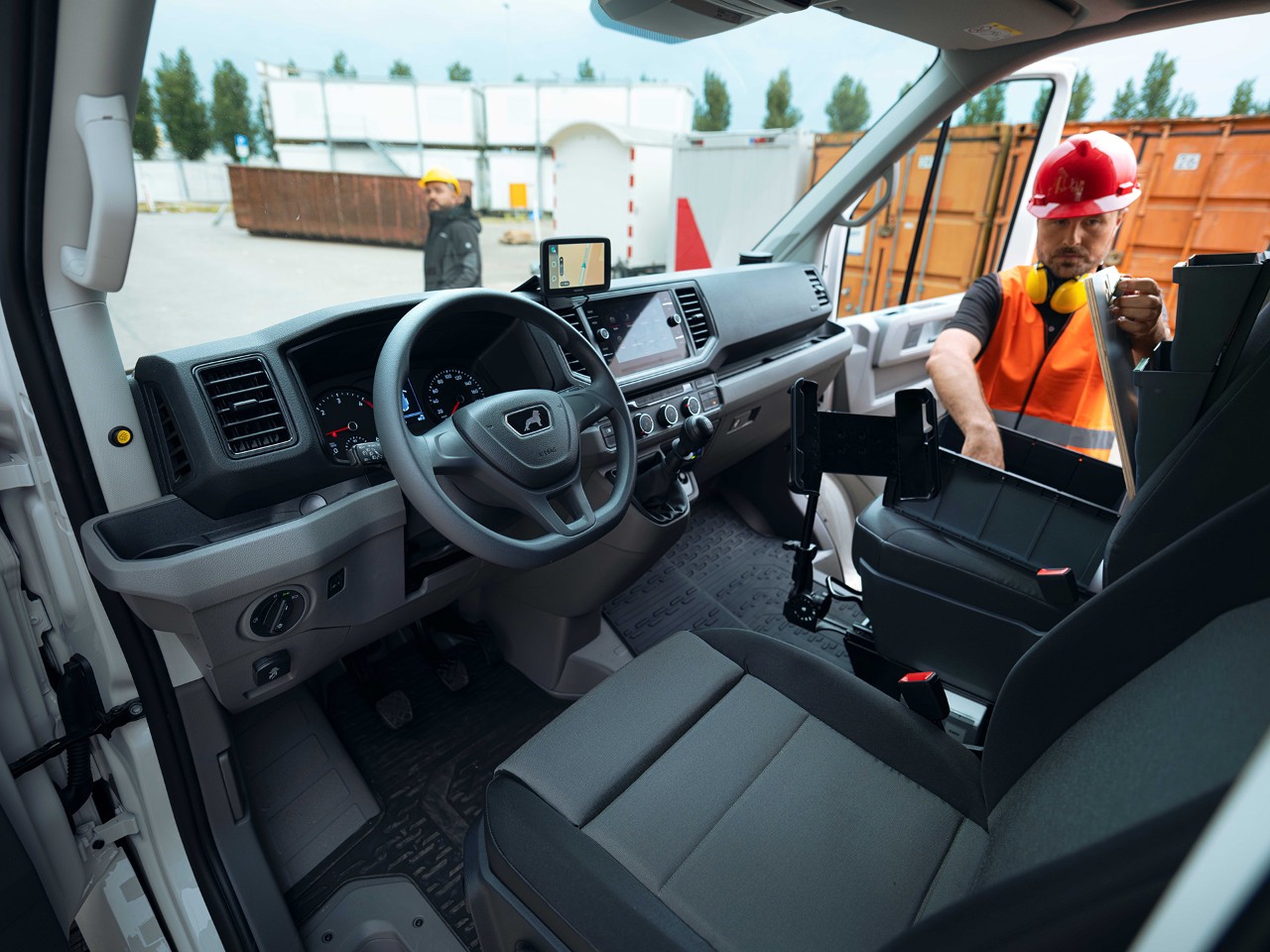A construction worker in an orange safety vest and red helmet is organising items inside the front seat area of a van. The interior is shown from the driver's perspective, and the worker is adjusting a container or toolbox. A person is visible in the background, and a large container is parked nearby.