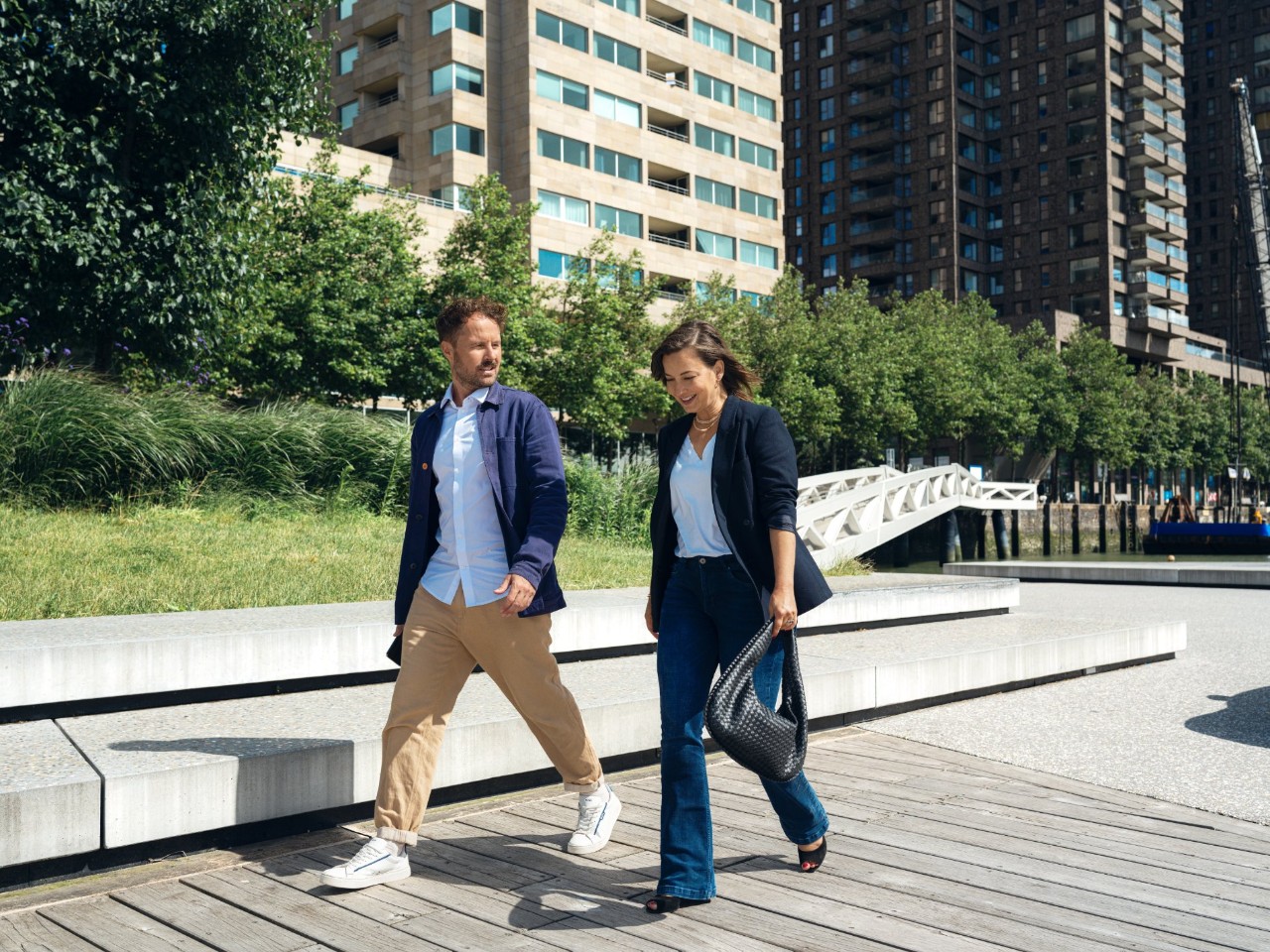 Man and woman walk down a pedestrain path together in the sunlight. There are skyscrappers behind them.