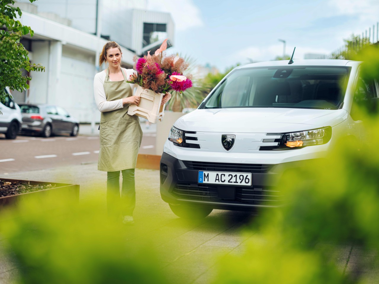 A florist wearing an apron carries a wooden crate filled with flowers, with a white delivery van parked beside her and greenery blurred in the foreground.