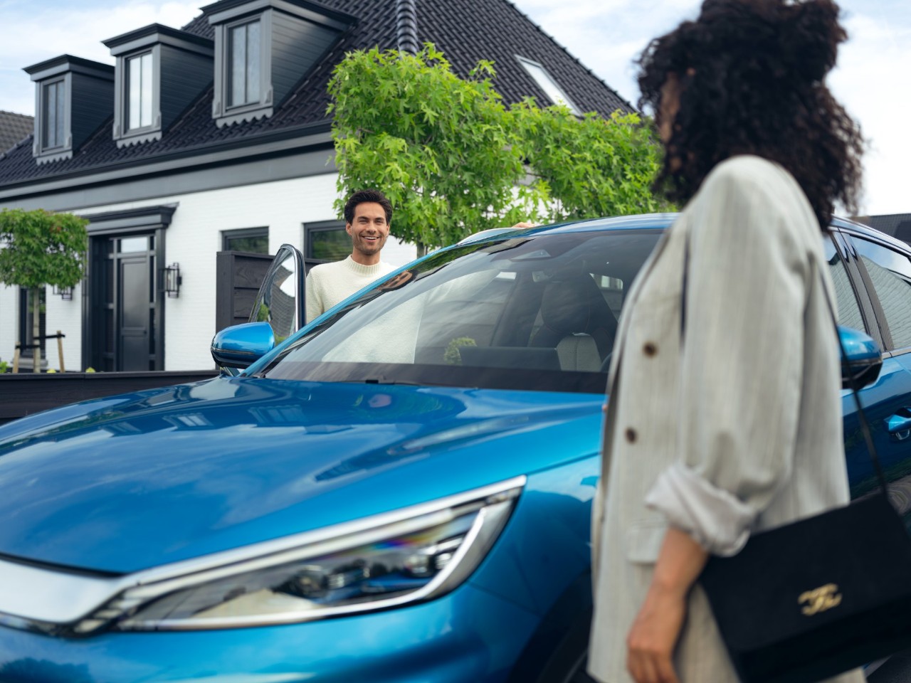 A man smiling and standing next to a blue car, holding the car door open. A woman in a striped blazer is walking towards the car. The background features a modern house with trees and greenery.