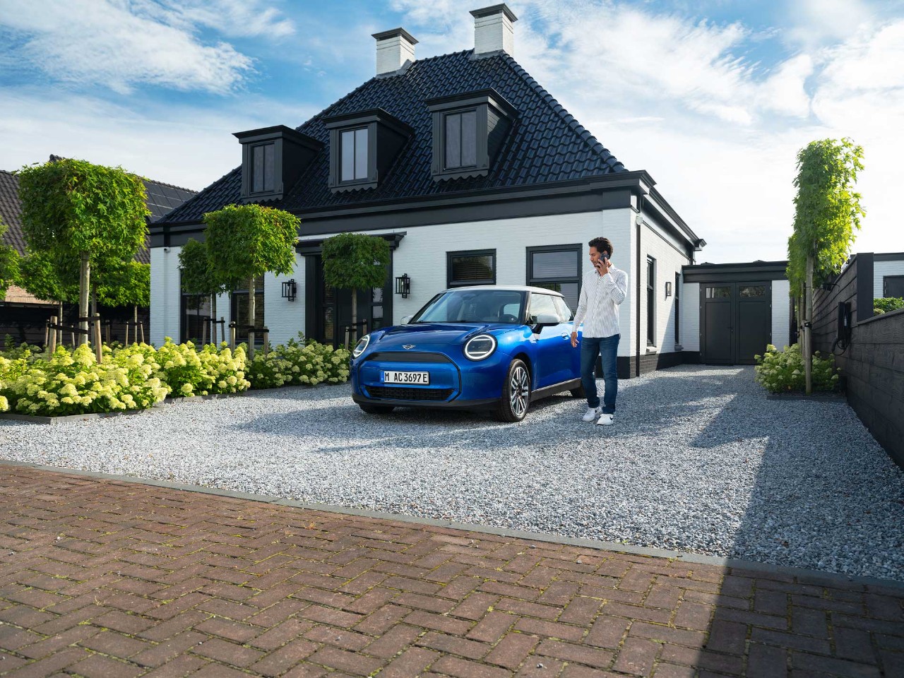 Man stands on the phone he walks next to a blue car parked on a gravel driveway. There is a modern house behind with trees and flowers.