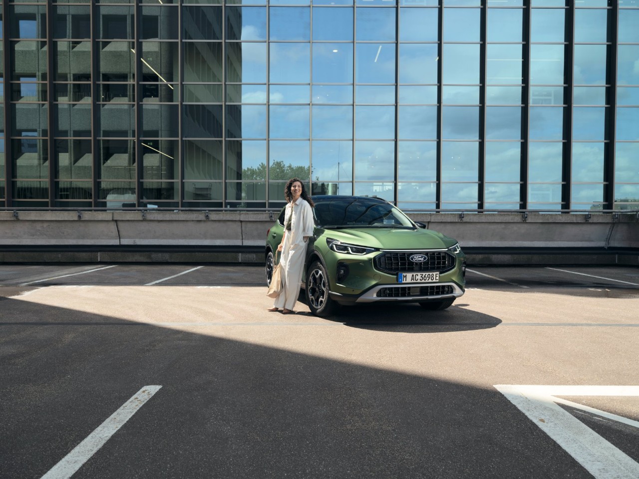 Woman leans against a green Ford vehicle in a parking lot in front of a glass facade.
