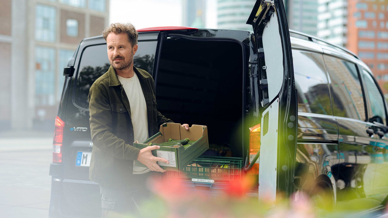 A man unloading crates of vegetables from the back of a black van. He is standing outside in a city environment with tall buildings in the background. The van's rear doors are open, and he is carrying a box of produce.