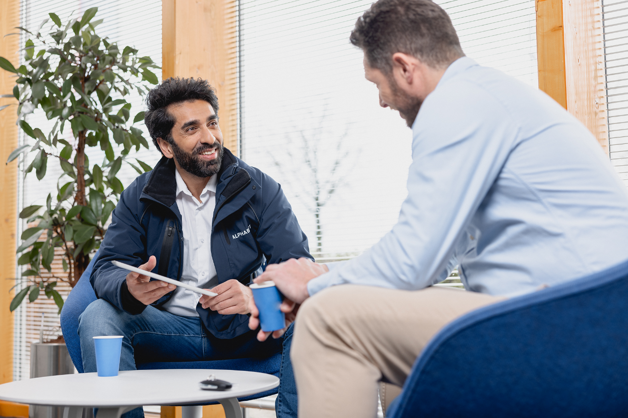 Two men having a conversation one holds a tablet the other has a coffee in their hand. There is a plant behind them.