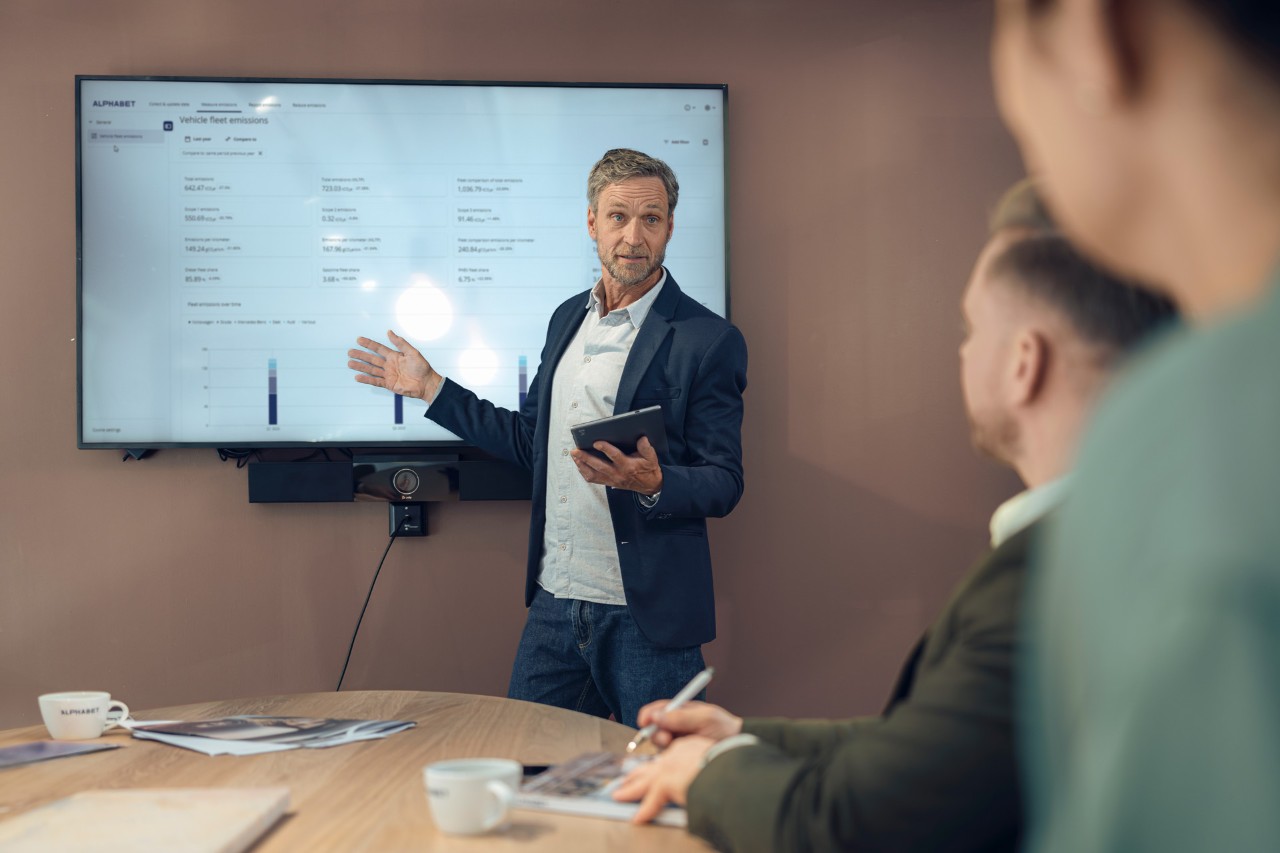 Man presenting sustainability information on a wall-mounted display to colleagues in a meeting room.