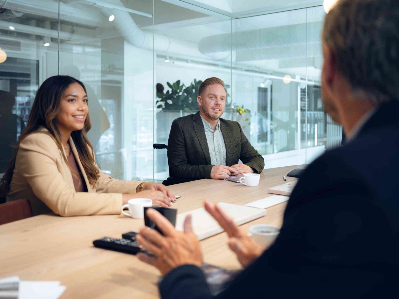Three people having a conversation at a wooden table in a modern office with glass walls.