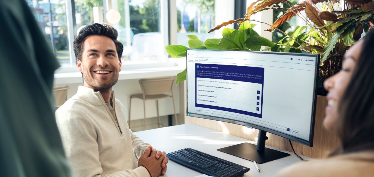 Person sitting at a desk in a modern office, working on a computer with the Alphabet website open, surrounded by green plants and large windows.