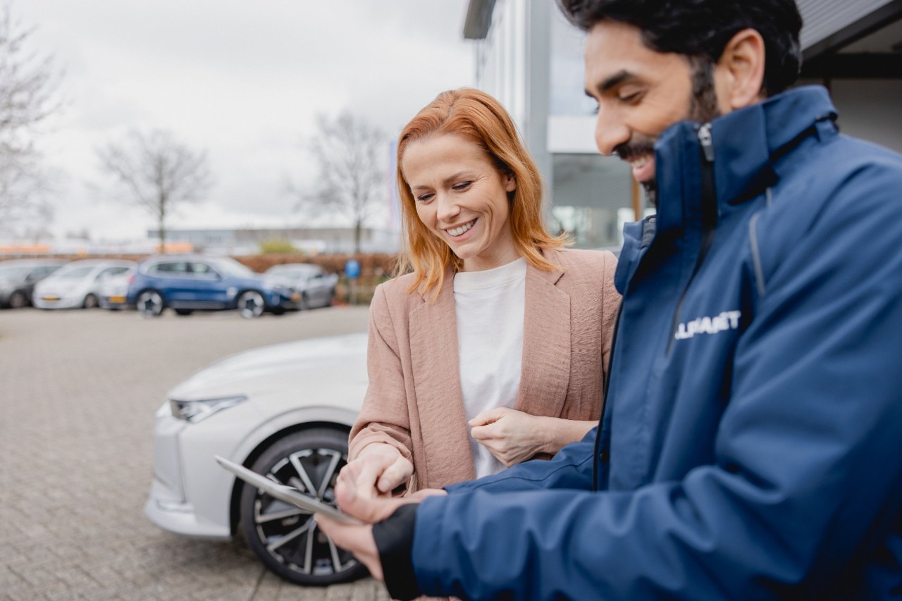Two people standing next to a white car, discussing information displayed on a tablet in an outdoor setting.