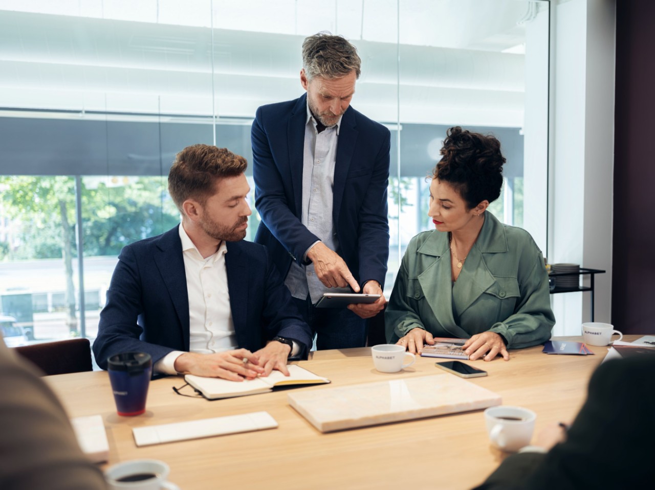 Three people having a meeting with a tablet in a modern office with natural light and a view of trees.