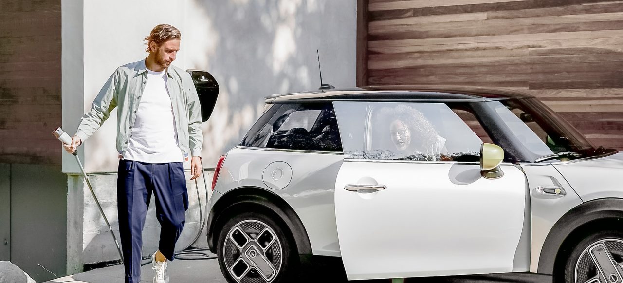 Hombre cargando su coche MINI y mujer saliendo del coche en su parking privado. Coche eléctrico blanco y chico rubio
