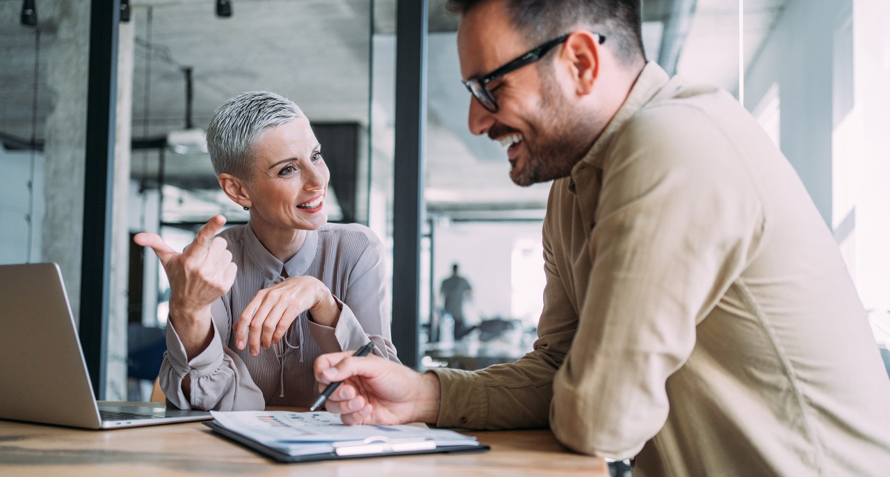 Shot of a two confident business persons sitting on a desk in the office and sharing ideas. Businessman and businesswoman in meeting using laptop and discussing business strategy. Business coworkers working together in the office.