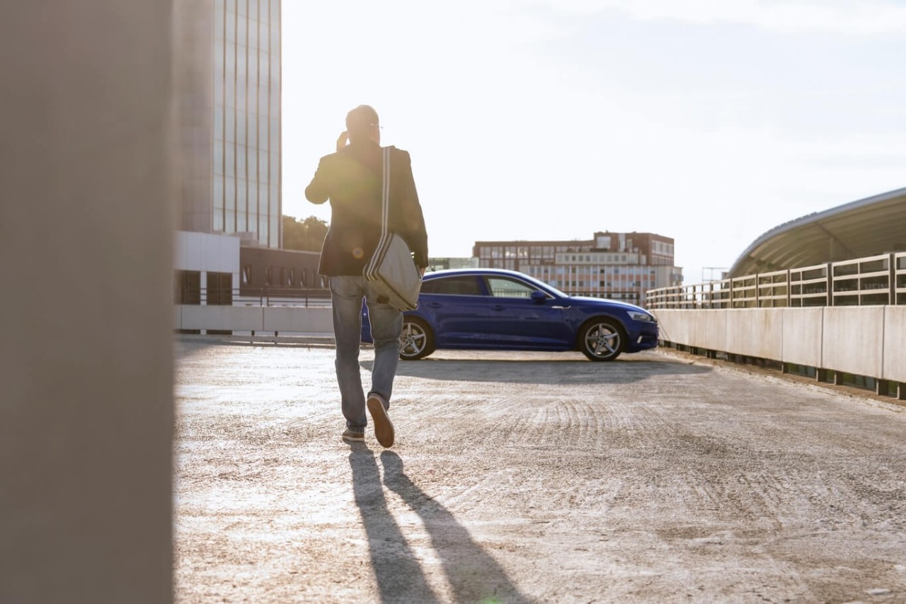 Hombre caminando hacia su coche aparcado en un parking al aire libre con el sol radiando. Chico alejandose con coche azul de fondo