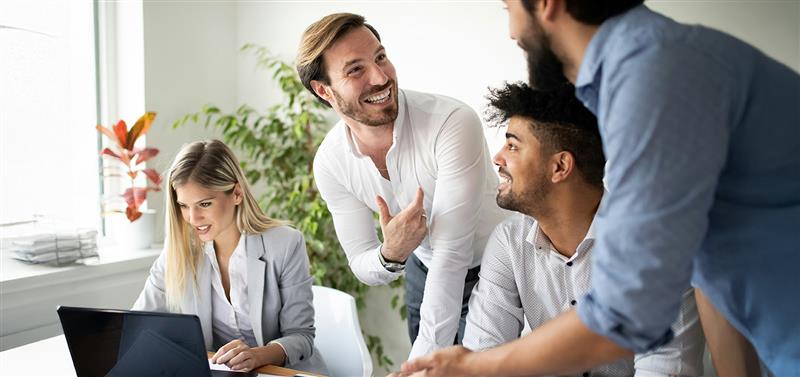 cuatro trabajadores colaborando con sonrisas, mirando el ordenador en una ofinica de colores claros