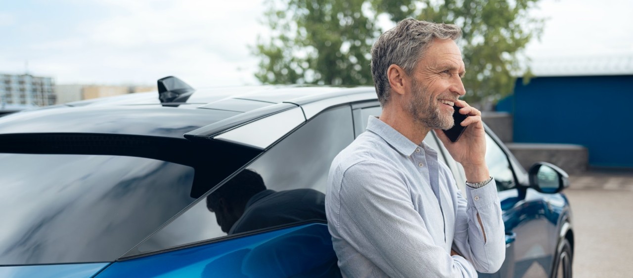 Man in a light shirt leans against a blue car while talking on a mobile phone in a parking lot.